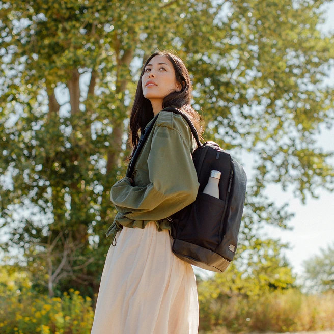 A woman wearing a backpack stands in front of a tree on a sunny day